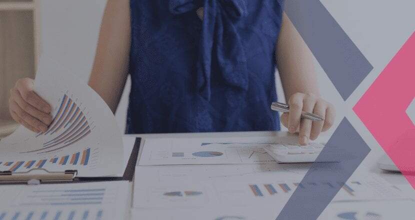 Mulher branca vestindo uma blusa sem manga na cor azul escura com um laço na gola da mesma cor, sentada a mesa, analisando gráficos e utilizando uma calculadora branca, fazendo cálculo da sua empresa.