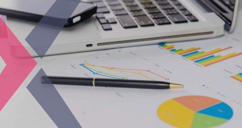 Uma mesa repleta de papéis com gráficos coloridos que representam o controle financeiro de um negócio, em cima dos papéis está uma caneta e uma laptop com um smartphone em cima deste.