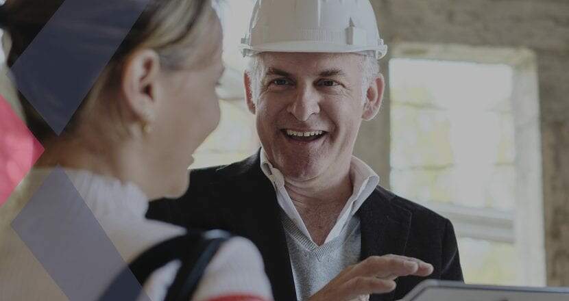 Uma mulher branca e de cabelos loiros presos conversando com um homem de 50 anos de cabelos brancos com um capacete de engenheiro sobre um projeto