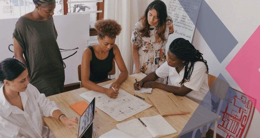 Grupo de 5 mulheres, 3 negras e 2 brancas, avaliando o balanço patrimonial da sua empresa em uma mesa de madeira cheia de papéis e um laptop