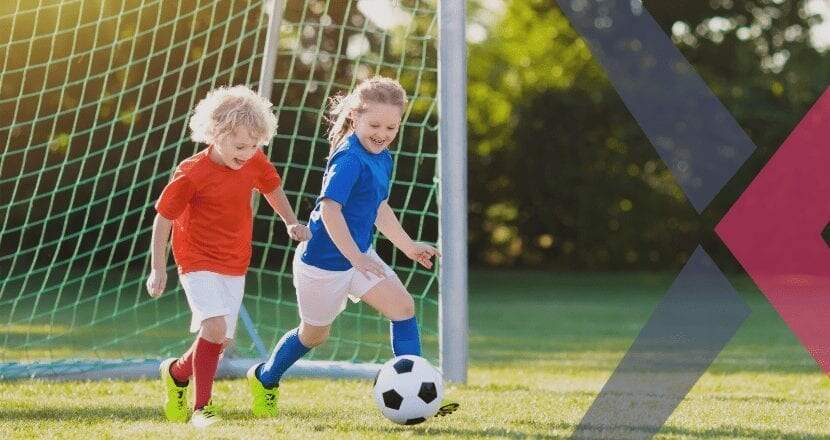 Duas crianças brancas, de cabelos loiros, jogando futebol. A menina está de camisa azul e short branco e o menino de blusa vermelha e short branco.