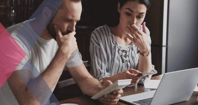 Um jovem casal caucasiano com cara de preocupados, ambos com a mão sobre o queixo, fazendo o cálculo de seus impostos em uma calculadora de cor branca e de frente para um laptop prata.