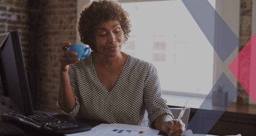 Mulher negra, em torno dos 50 anos, com cabelos crespos tomando café em uma xícara azul e fazendo o planejamento do seu MEI
